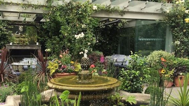 Een buitenplaats met een tuin, een fontein, plantenbakken en een eettafel.
