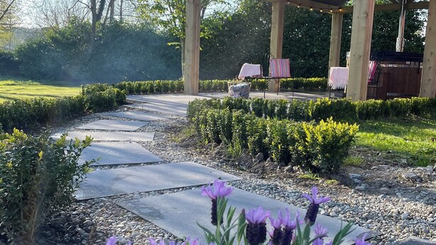 Ein überdachter Außenbereich mit Pergola, umgeben von einem angelegten Garten mit Kiesweg und violetten Blüten.