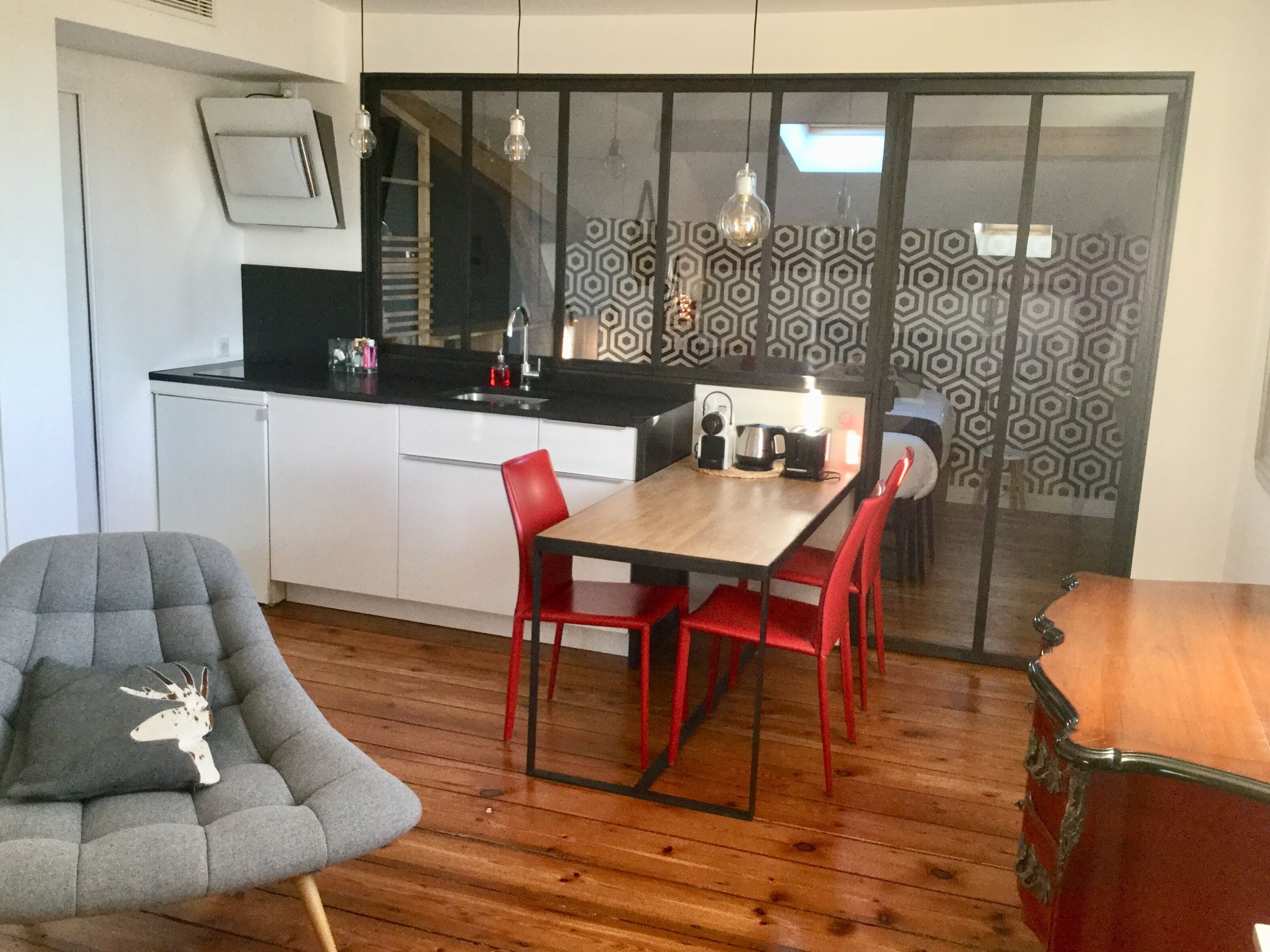 Un espace de vie compact comprenant un coin cuisine, un coin repas avec une table en bois et des chaises rouges, ainsi qu’un fauteuil gris agrémenté d’un coussin décoratif.