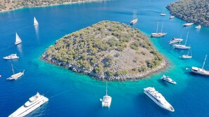 An aerial view of a small island surrounded by clear blue waters with multiple yachts anchored around it.