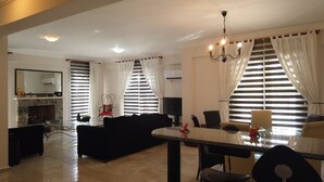 A modern living room with a fireplace, a dining area with a glass table, and large windows with curtains. - Calis Beach - Beautiful 4 bedroom villa with private pool and WiFi  (Fethiye)