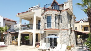 A two-story house with a stone facade, a balcony with metal railings, and a covered outdoor seating area with white lounge chairs. - Calis Beach - Beautiful 4 bedroom villa with private pool and WiFi  (Fethiye)
