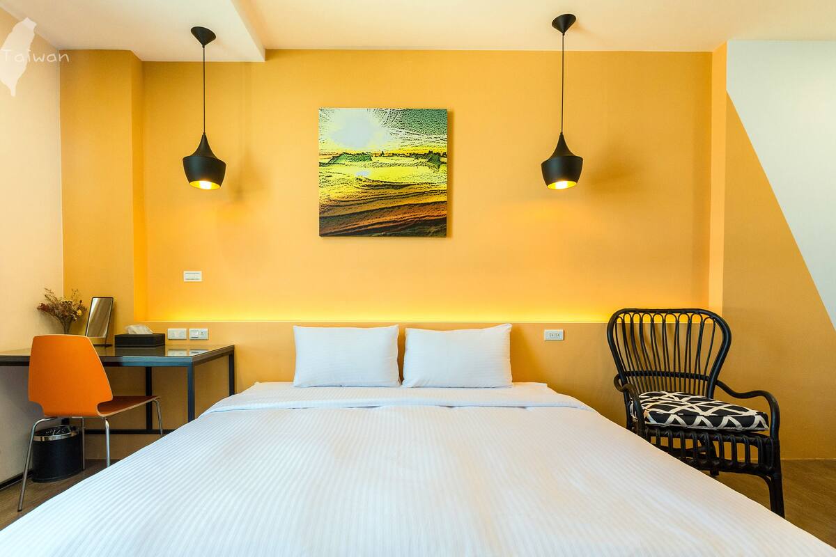 Habitación de hotel con una cama grande, dos lámparas colgantes, un escritorio con una silla y un cuadro en la pared.
