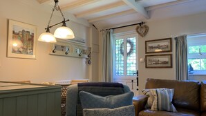 A cozy living room with a brown leather sofa, a green armchair, and a blue patterned armchair. There are two wall-mounted lamps, a framed picture of a cityscape, and a window with curtains. - Riverside cottage in a beautiful garden in Lincolnshire. (Boston)