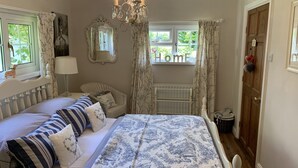 A bedroom with a large bed, a chair, a chandelier, a window with curtains, a mirror, and a door. - Riverside cottage in a beautiful garden in Lincolnshire. (Boston)