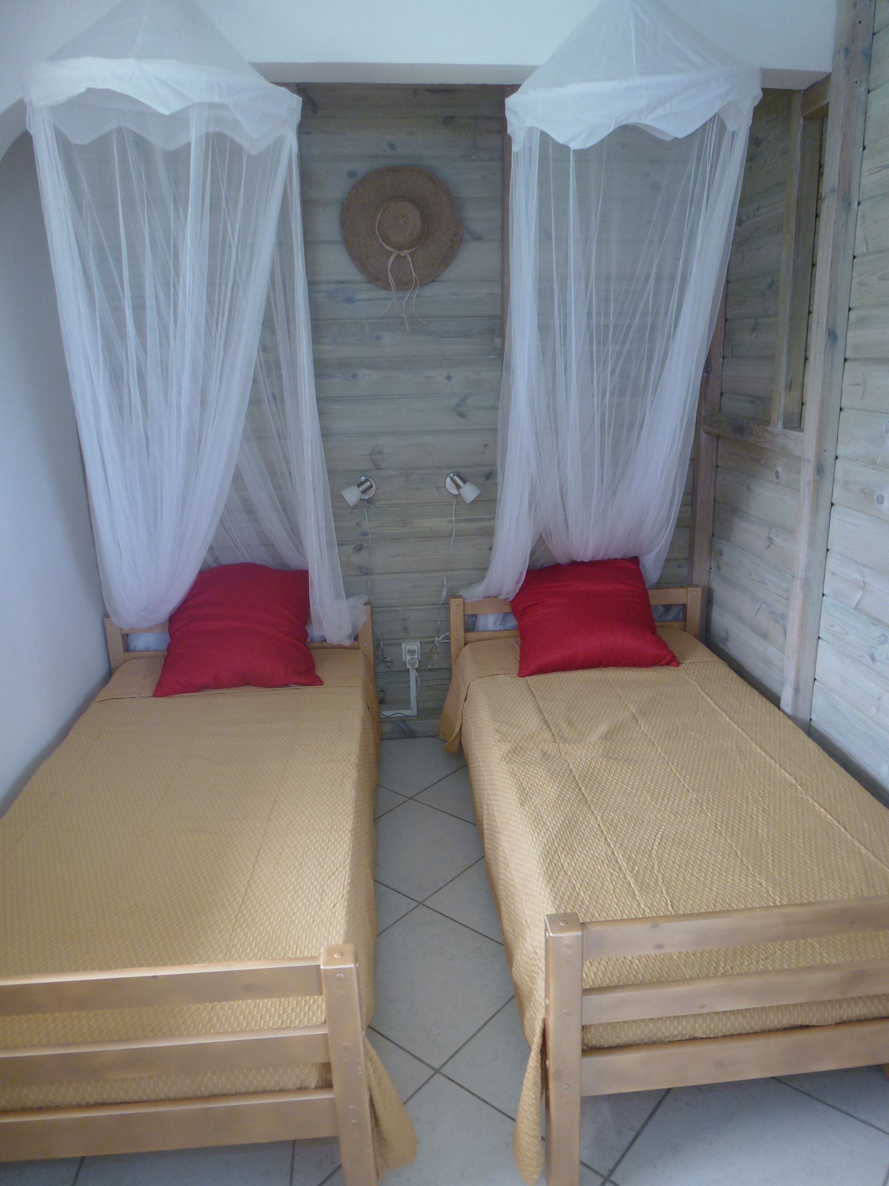 Deux lits simples avec des baldaquins blancs, des oreillers rouges et une tête de lit en bois.