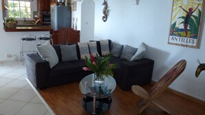 A living room with a black sofa, a glass coffee table, and a wooden chair. There is a kitchen in the background with a refrigerator and a window. - The KREYOL LODGES presents its SUITE KAYOU ... view sea and mountain (DESHAIES)