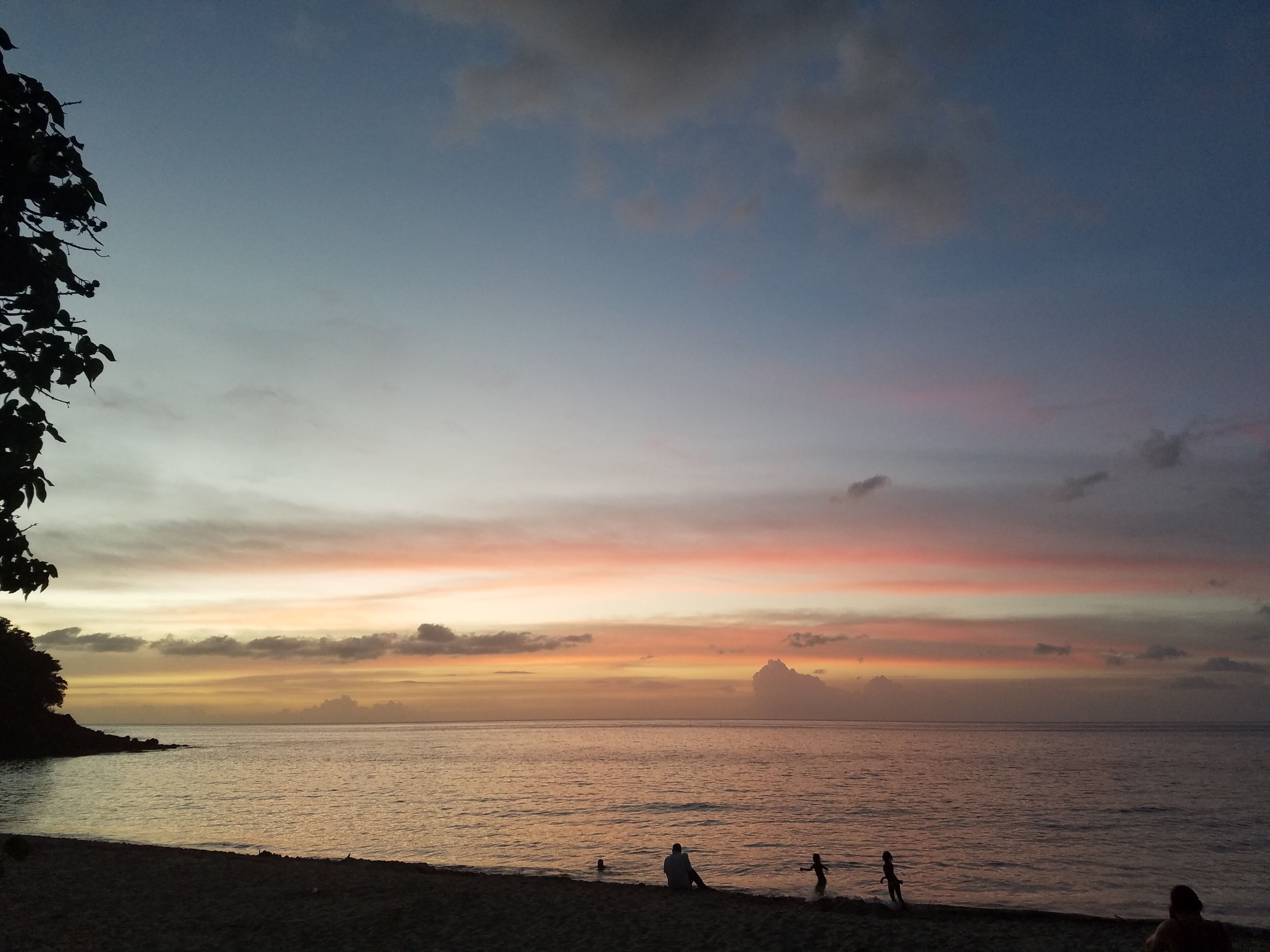 Eine Strandansicht mit Sonnenuntergang über dem Meer, Menschen sitzen und spielen nahe am Wasser.