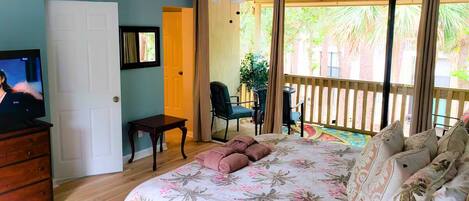 A bedroom with a bed, a TV, a ceiling fan, a door, a chair, and a balcony with a view of palm trees.