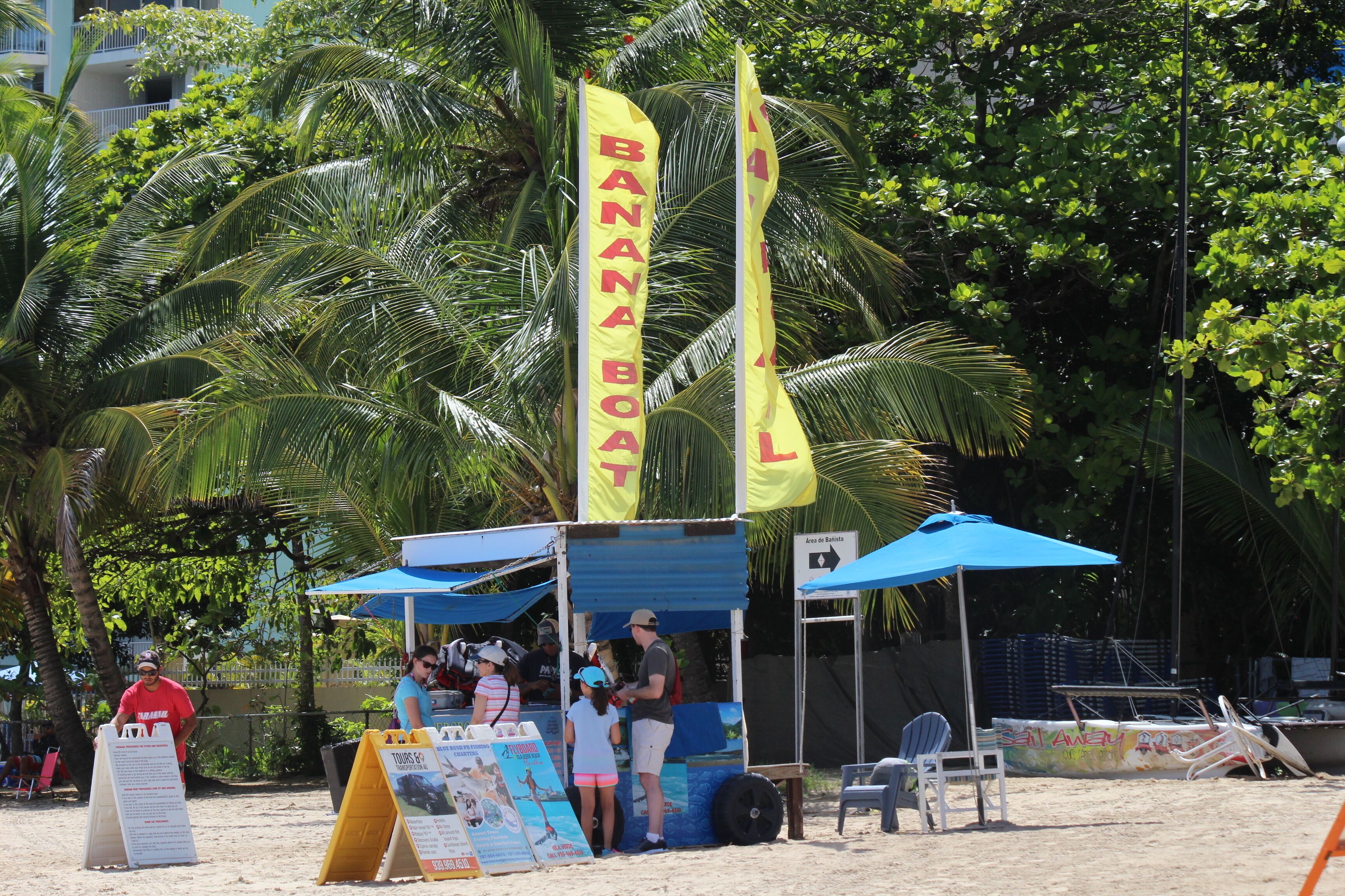 A beachside food cart with a yellow flag labeled 'BANANA BOAT' and a sign indicating 'Area de Banister.'