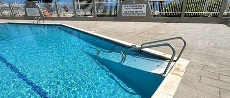 Ein klares Schwimmbecken mit Rettungsschwimmer-Schild, einer gefliesten Liegefläche und Palmen im Hintergrund.