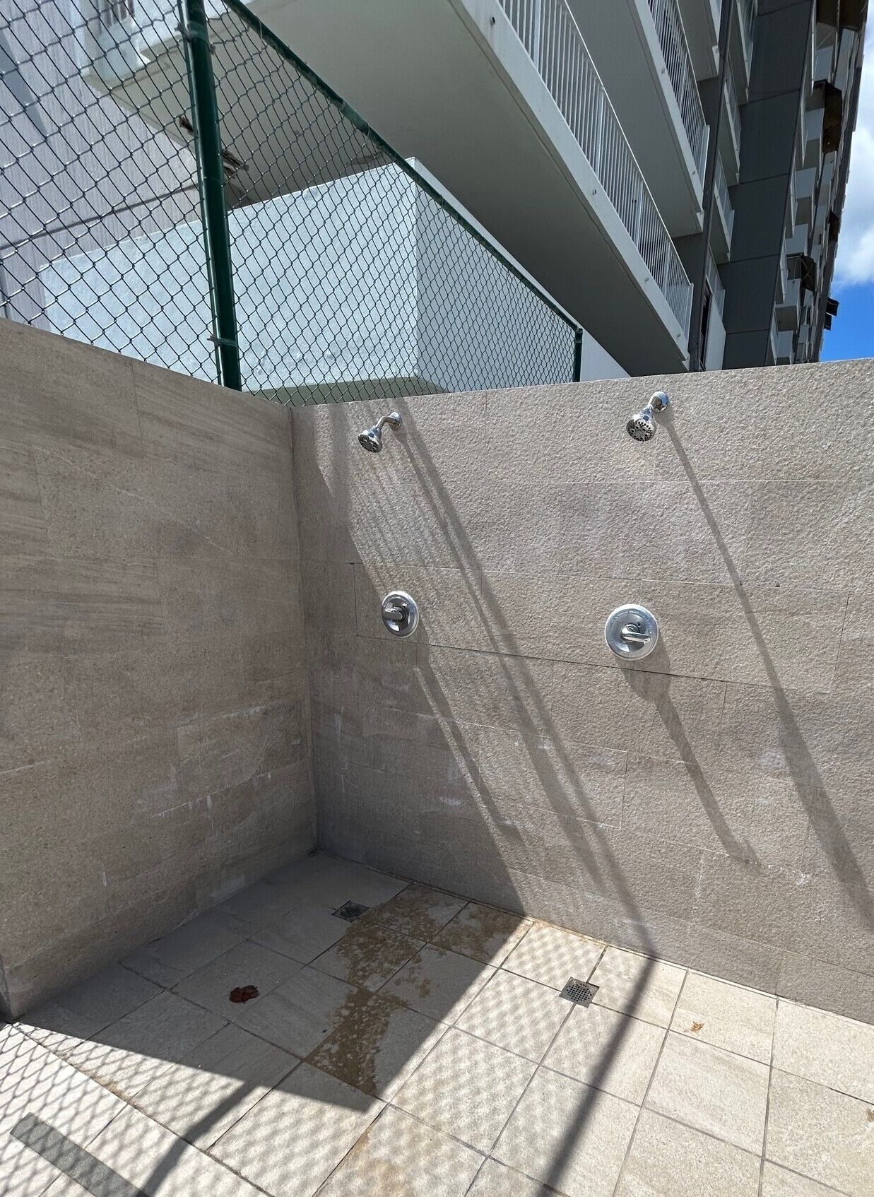 A concrete outdoor shower area with four chrome fixtures, surrounded by a chain-link fence and a building in the background.