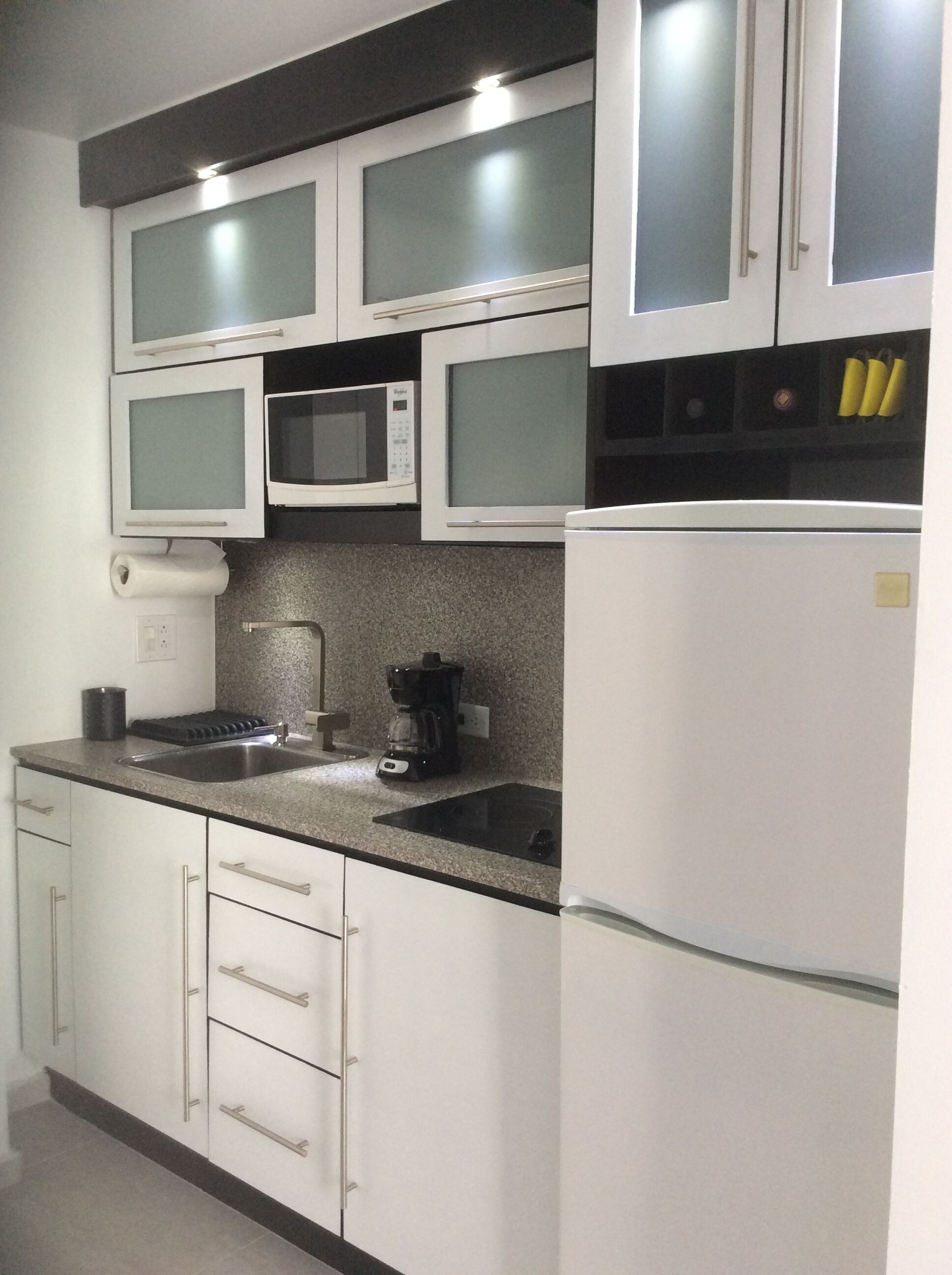 A kitchen with white cabinets, a stainless steel sink, a microwave, a coffee maker, and a refrigerator.