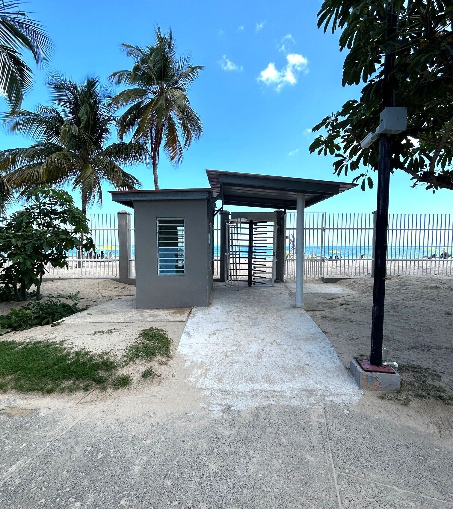A small building with a covered entrance, a concrete pathway leading to it, and a beach in the background.