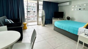 Ein modernes Hotelzimmer mit Bett, Fernseher, Schreibtisch und Stuhl. Ein Balkon bietet Blick auf die Stadt.