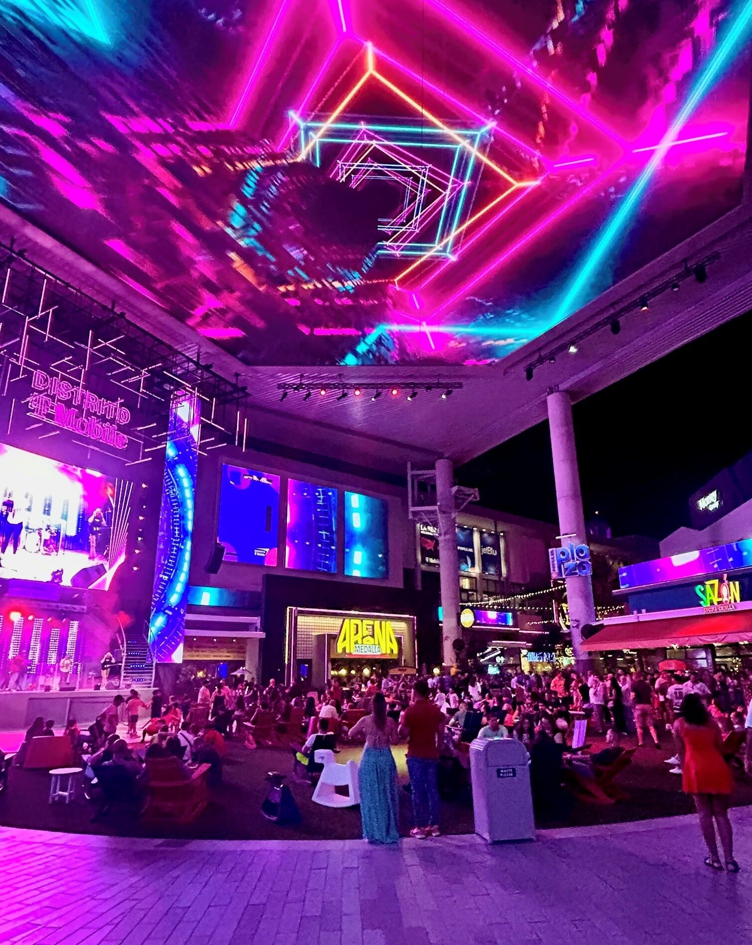 Un espacio exterior vibrante con un escenario y asientos para un evento, rodeado de estructuras e indicaciones iluminadas.