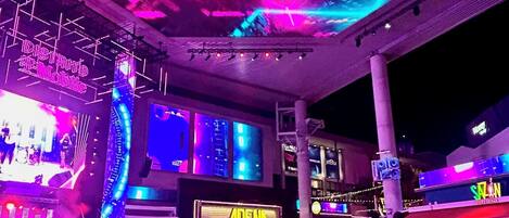 Un espacio exterior vibrante con un escenario y asientos para un evento, rodeado de estructuras e indicaciones iluminadas.