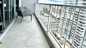 Ein Balkon mit Glasdoor, einem kleinen Tisch und einem Stuhl. An der Wand hängt eine Uhr und es bietet sich ein Blick auf eine Skyline mit Hochhäusern.
