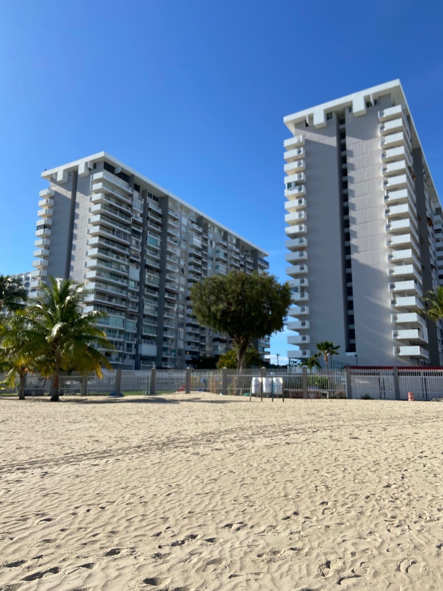 A beachfront hotel with multiple floors, a sandy beach in the foreground, and a clear blue sky.