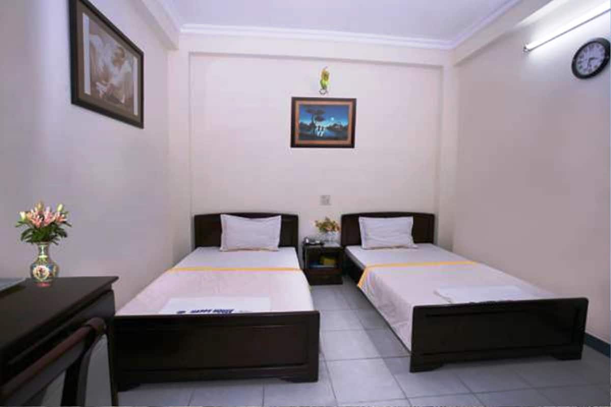 Une chambre d’hôtel avec deux lits simples, un bureau, une chaise, un vase avec des fleurs, une horloge au mur et un tableau encadré.