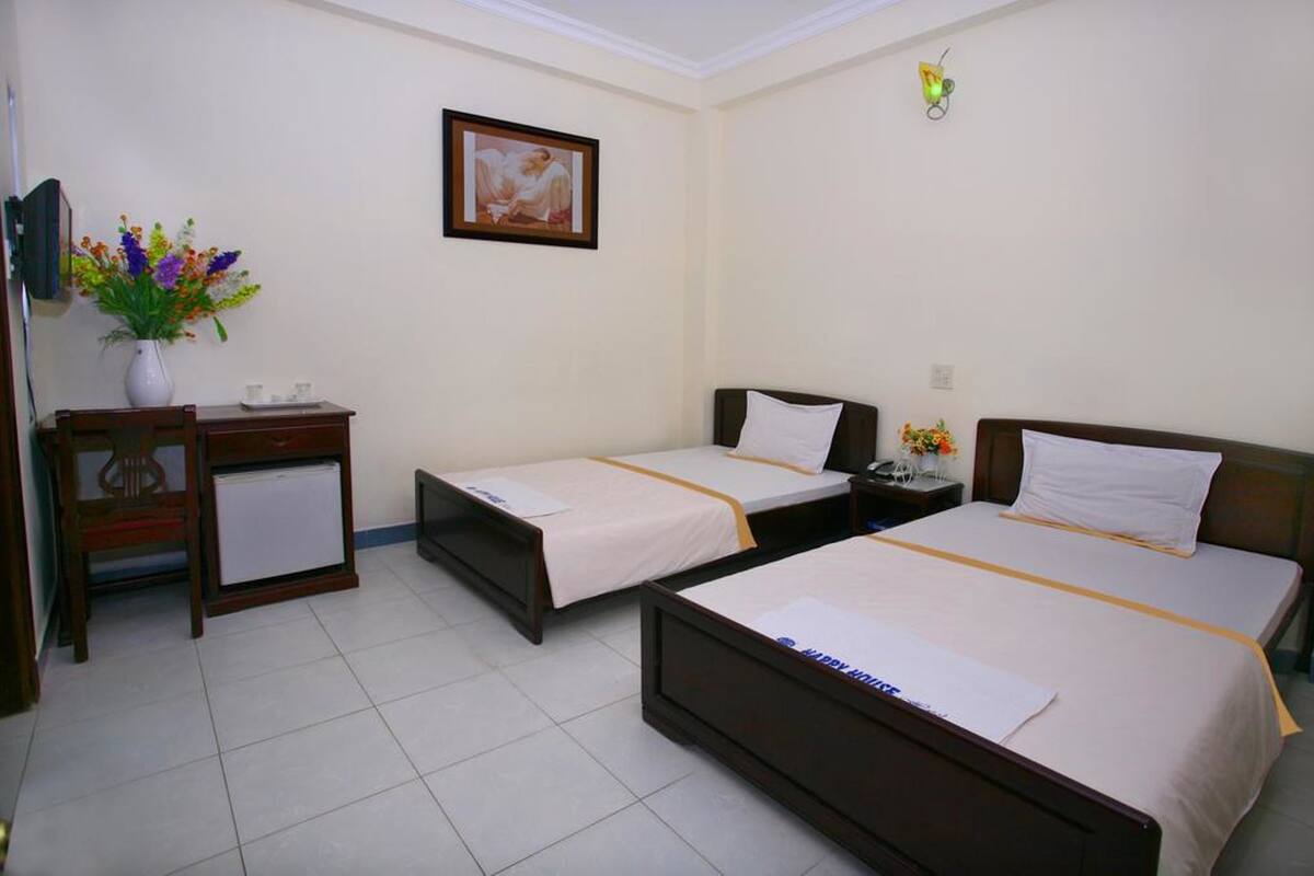 Une chambre d’hôtel avec deux lits simples, un petit bureau avec une chaise, un réfrigérateur, un vase avec des fleurs et un tableau encadré au mur.