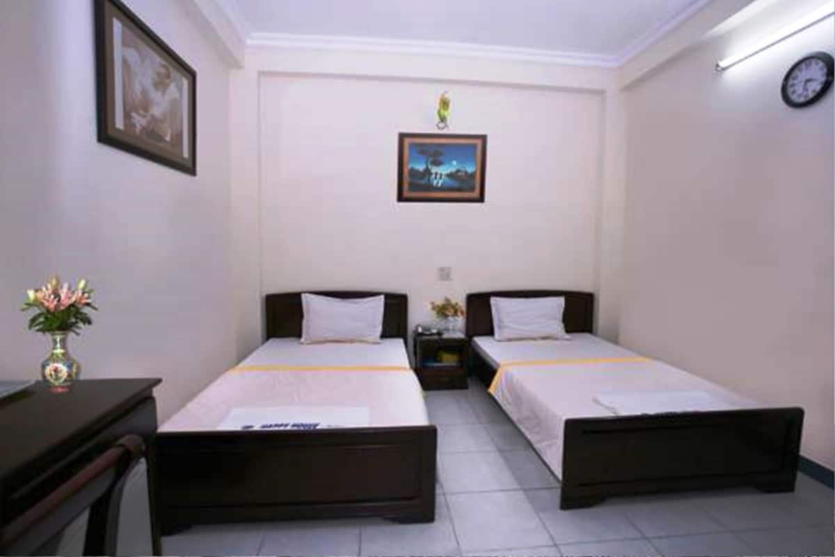 Une chambre d’hôtel avec deux lits simples, un bureau, une chaise, une petite table avec un vase de fleurs et une horloge au mur.