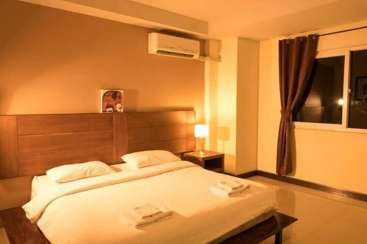 Habitación de hotel con cama, mesita de noche con lámpara, unidad de aire acondicionado y ventana con cortinas.