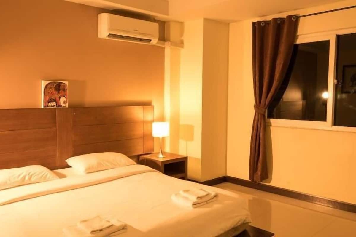 Quarto de hotel com cama, mesa de cabeceira, luminária, aparelho de ar condicionado e janela com cortinas.