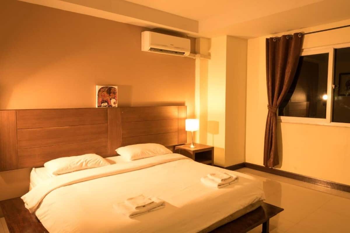Quarto de hotel com uma cama, mesa de cabeceira com abajur, aparelho de ar condicionado e janela com cortinas.