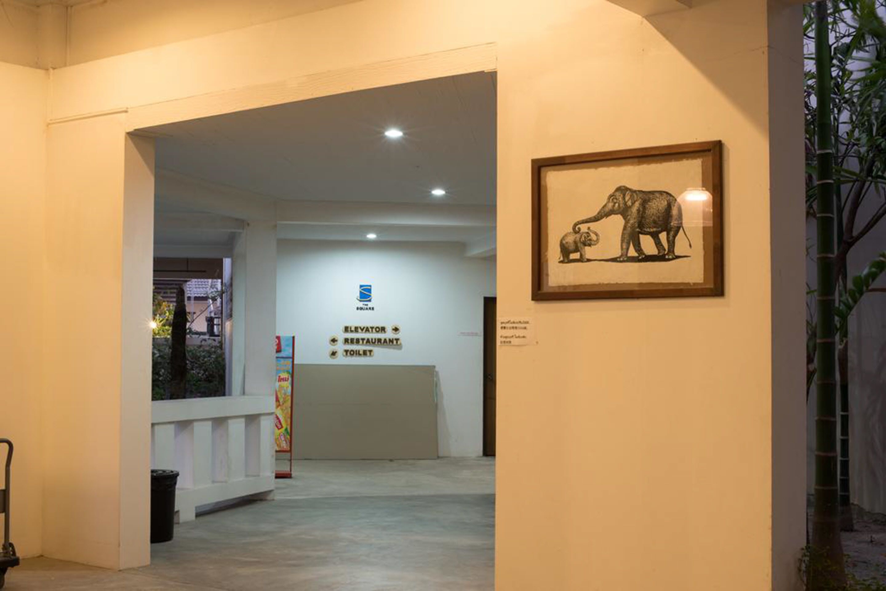 A hallway with a reception desk, a framed elephant artwork, and a sign indicating an elevator, restaurant, and toilet.
