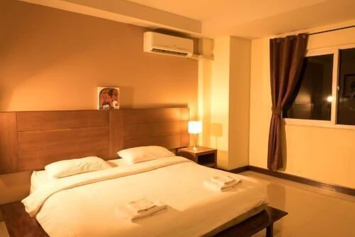 Quarto de hotel com cama, mesa de cabeceira, ar condicionado, janela com cortinas e um quadro pendurado na parede.