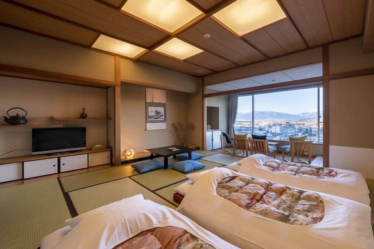A traditional Japanese-style room with tatami flooring, two futons, a television, a desk, and a view of a cityscape.