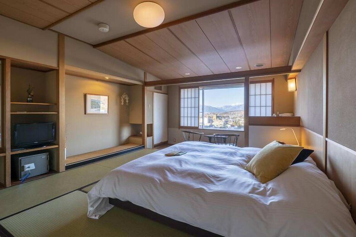 A traditional Japanese-style bedroom with tatami flooring, a bed, a television, and a view of a cityscape.