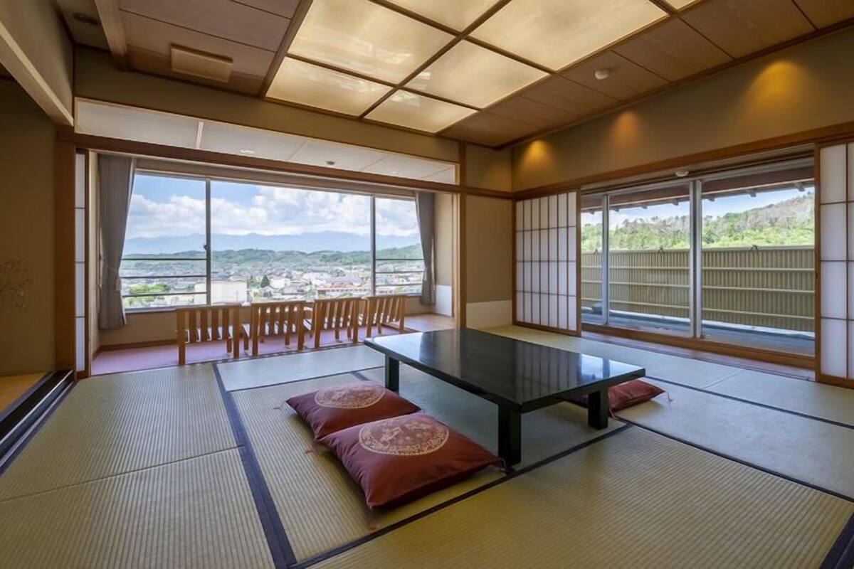A tatami mat room with a low table, cushions, and large windows offering a view of a town and mountains.