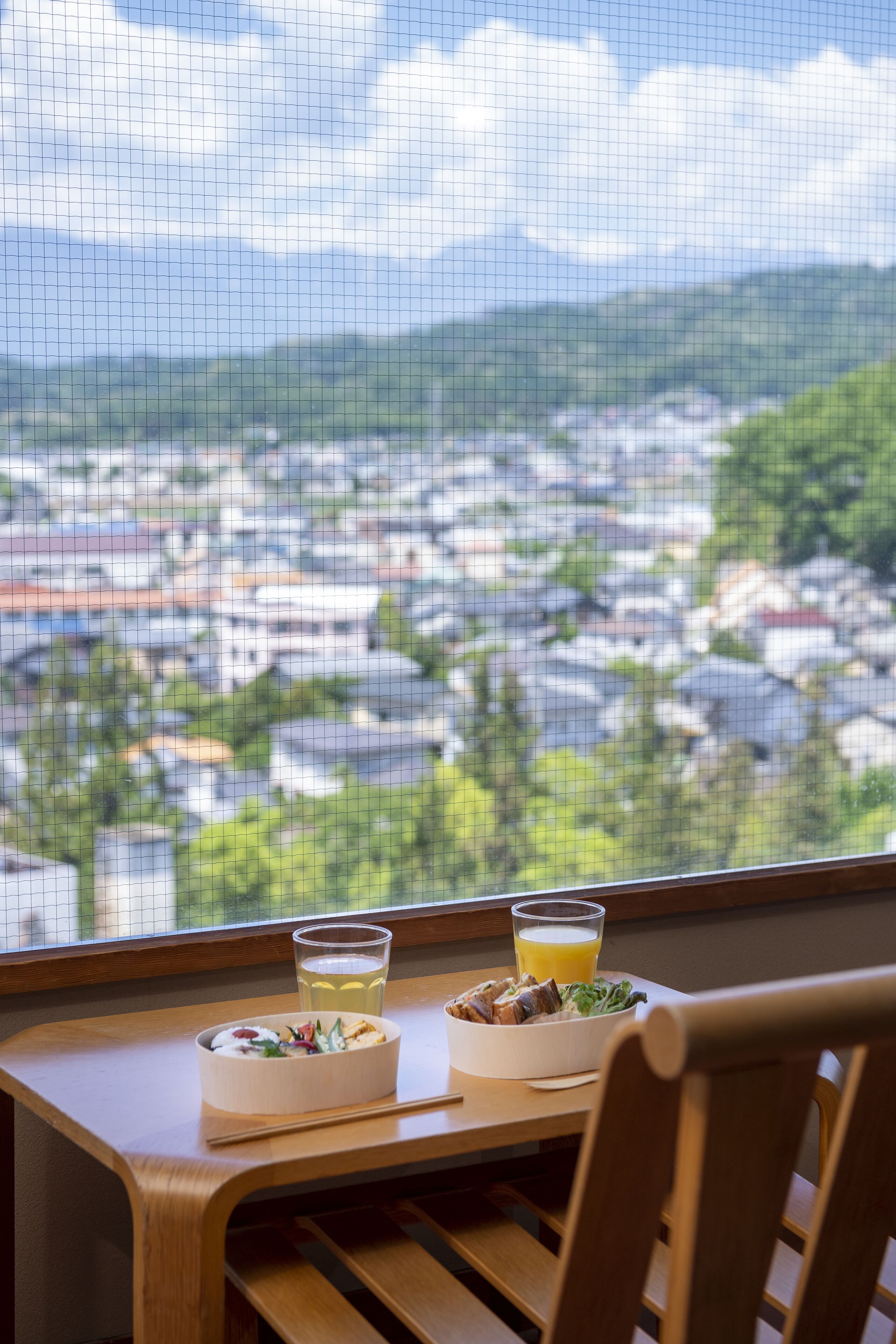 A dining table with two bowls of food and two glasses of beverages, set against a window overlooking a cityscape.