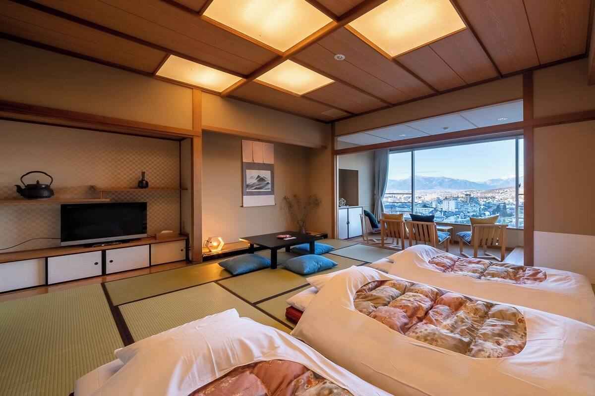 A traditional Japanese room with tatami flooring, a television, a desk, and a view of a cityscape.