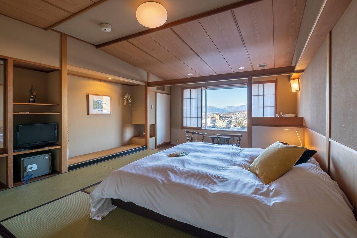 A traditional Japanese-style hotel room with tatami flooring, a bed with pillows, a television, and a view of a cityscape through large windows.
