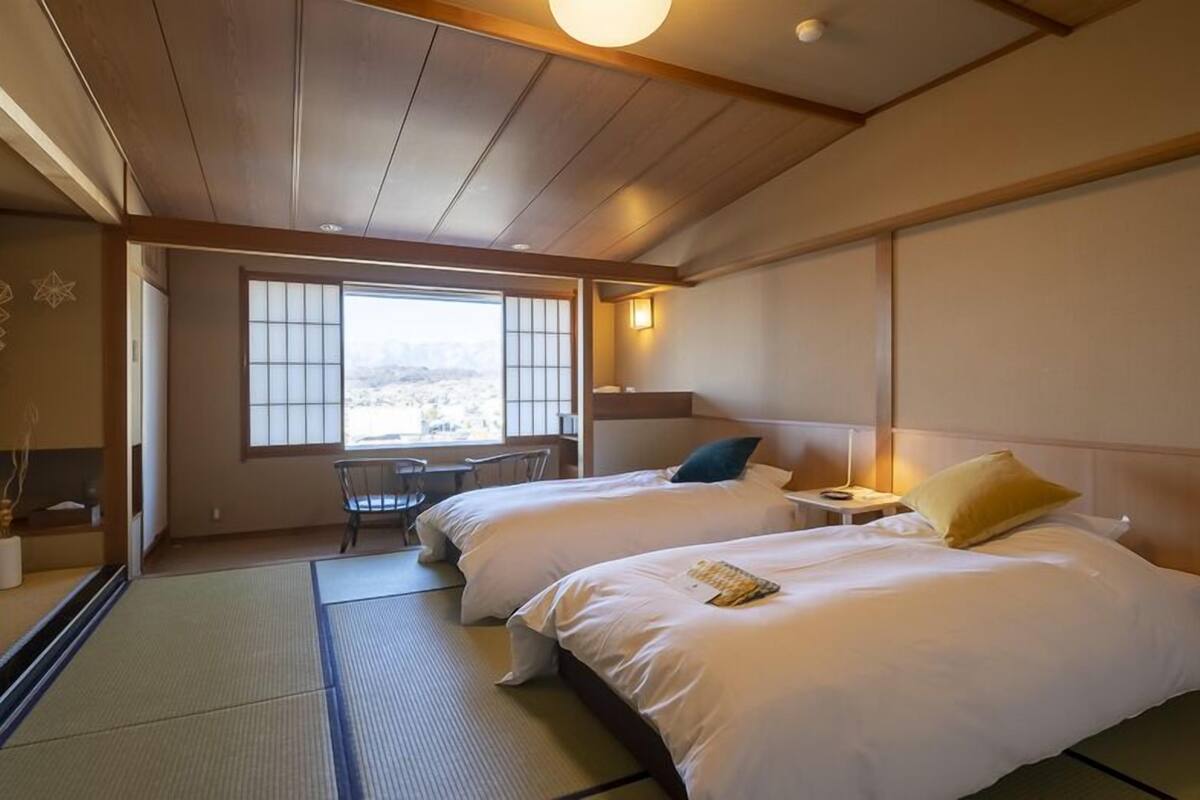 A traditional Japanese-style hotel room with tatami flooring, two beds, a desk, and a view of a mountainous landscape.