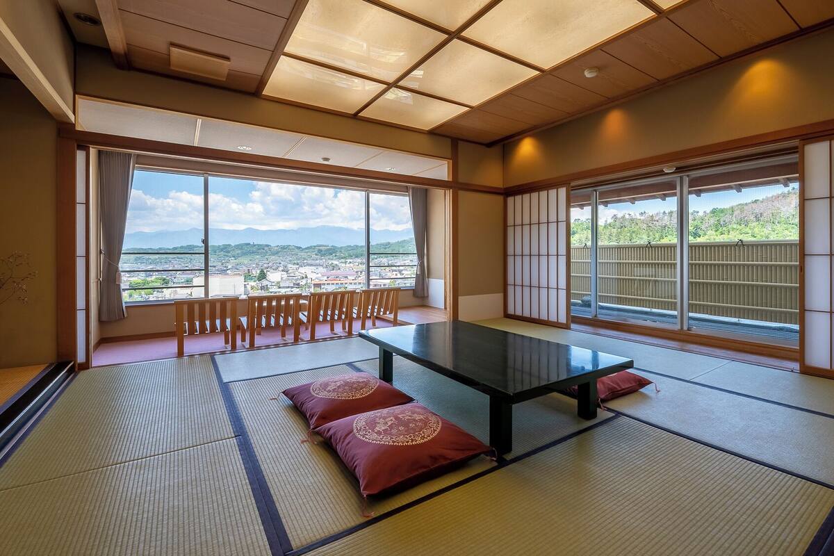 A tatami mat room with a low table and cushions, large windows with a view of a cityscape and mountains, and sliding doors.