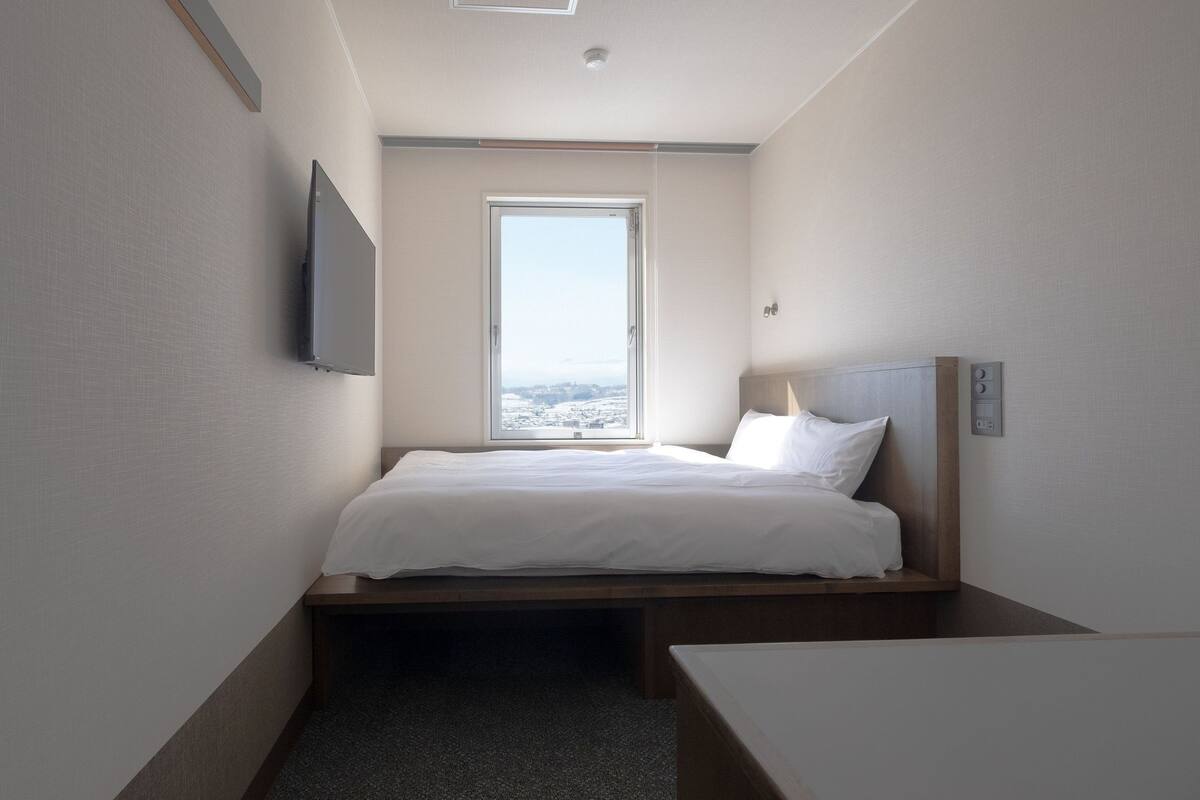 A small, modern hotel room with a bed, a TV, a window, and a bedside table.