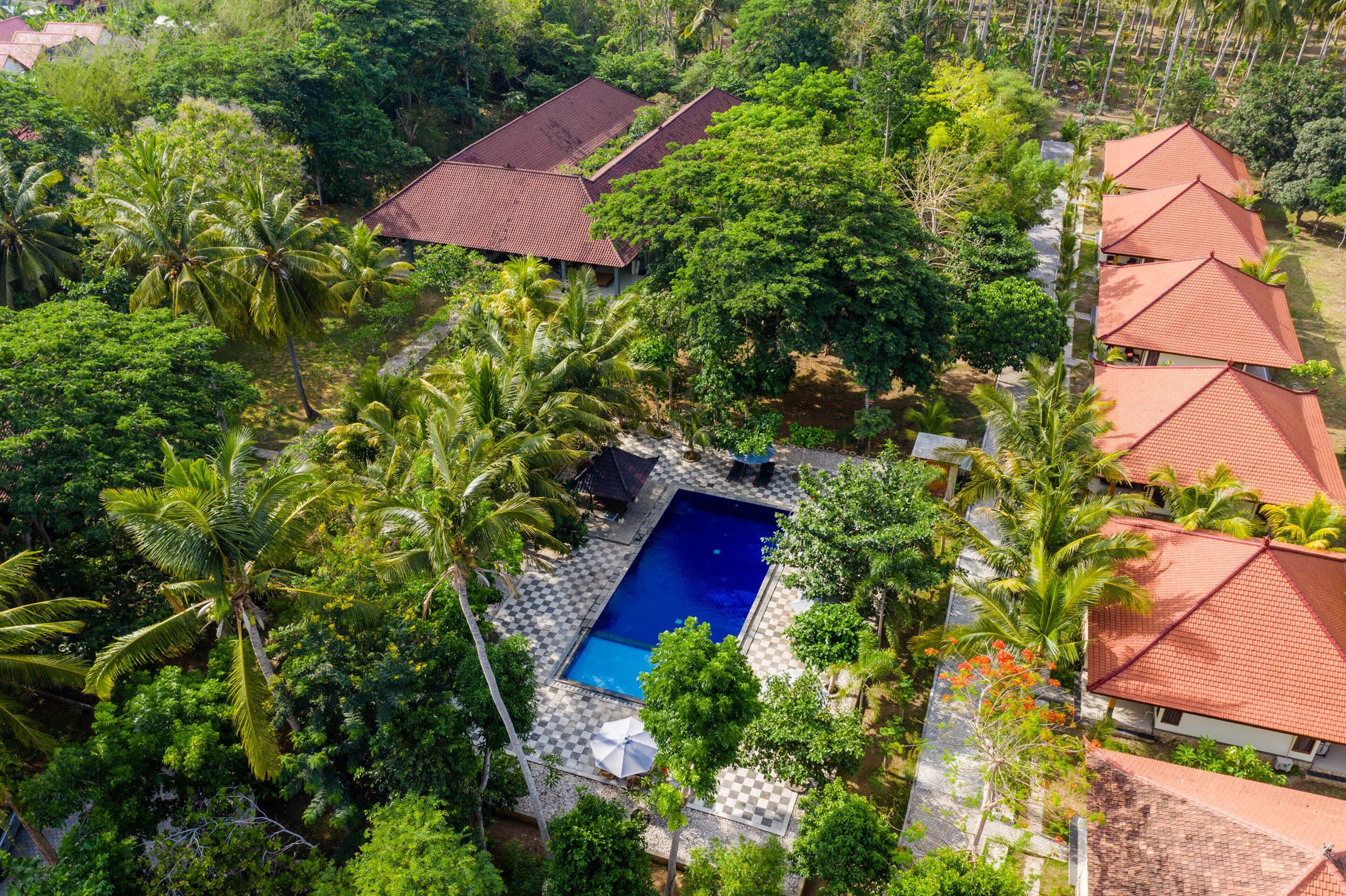 An aerial view of a resort with a swimming pool surrounded by palm trees and red-roofed buildings.