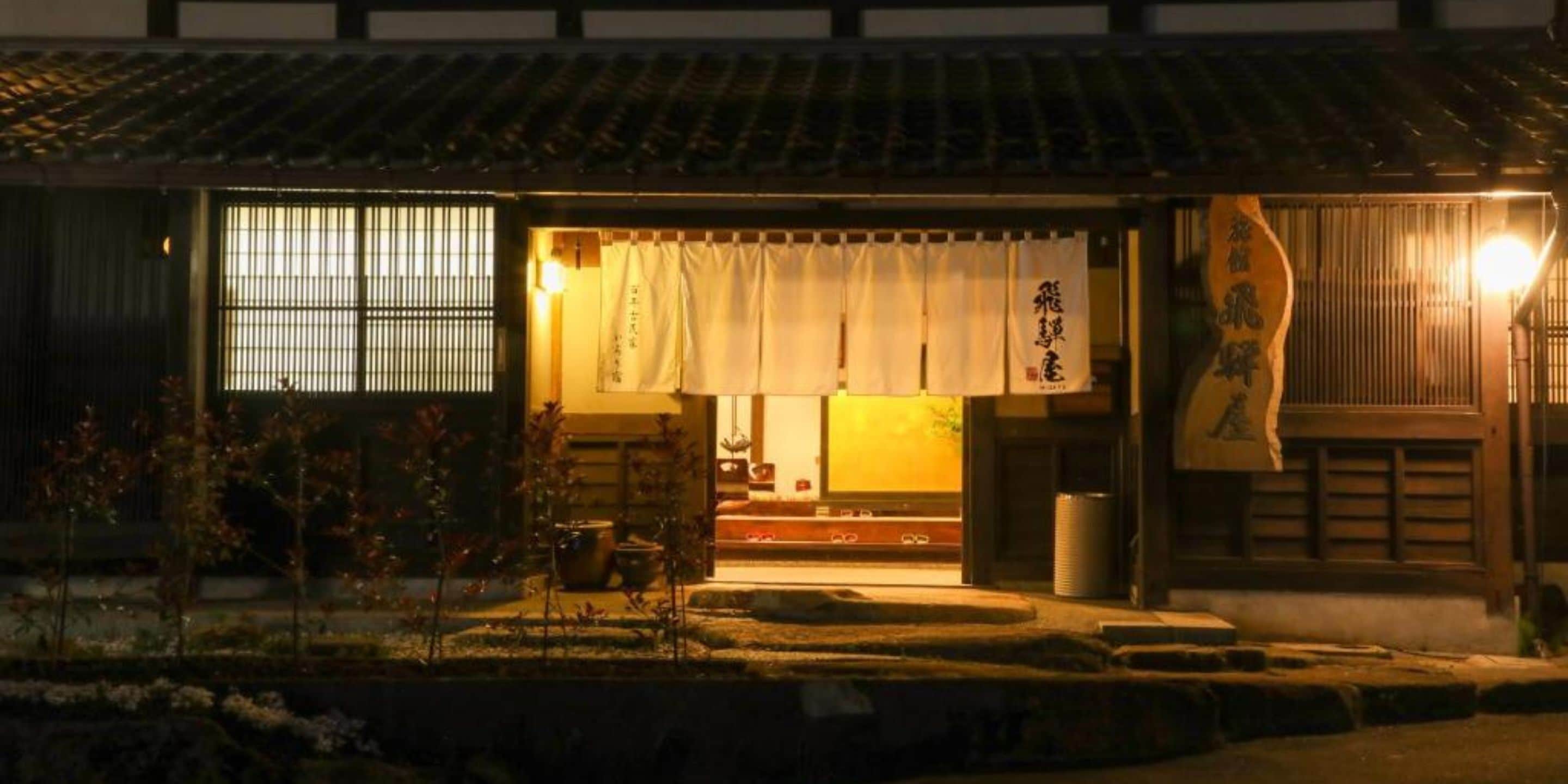 A traditional Japanese building with a wooden exterior, a tiled roof, and a large entrance with sliding doors. There is a sign with Japanese characters on the right side of the entrance.