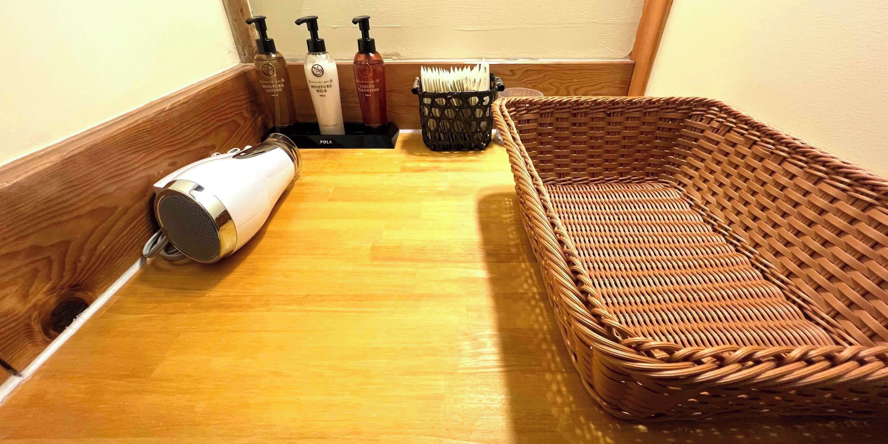 A room with a wooden floor, a white hairdryer, a wicker basket, and bottles of lotion and liquid soap.