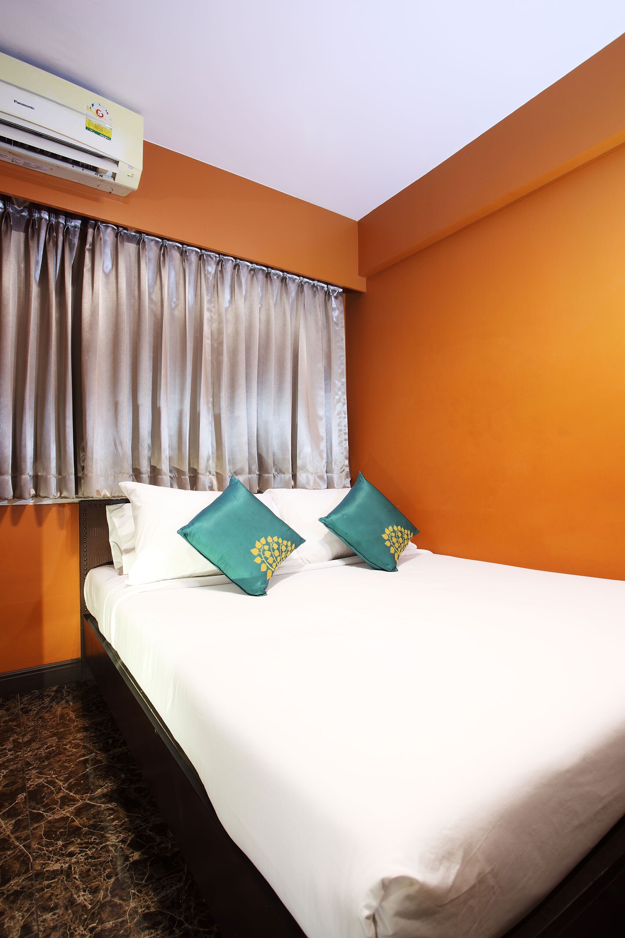 A hotel room with a bed, white bedding, two teal pillows with yellow designs, an air conditioning unit, and a window with curtains.