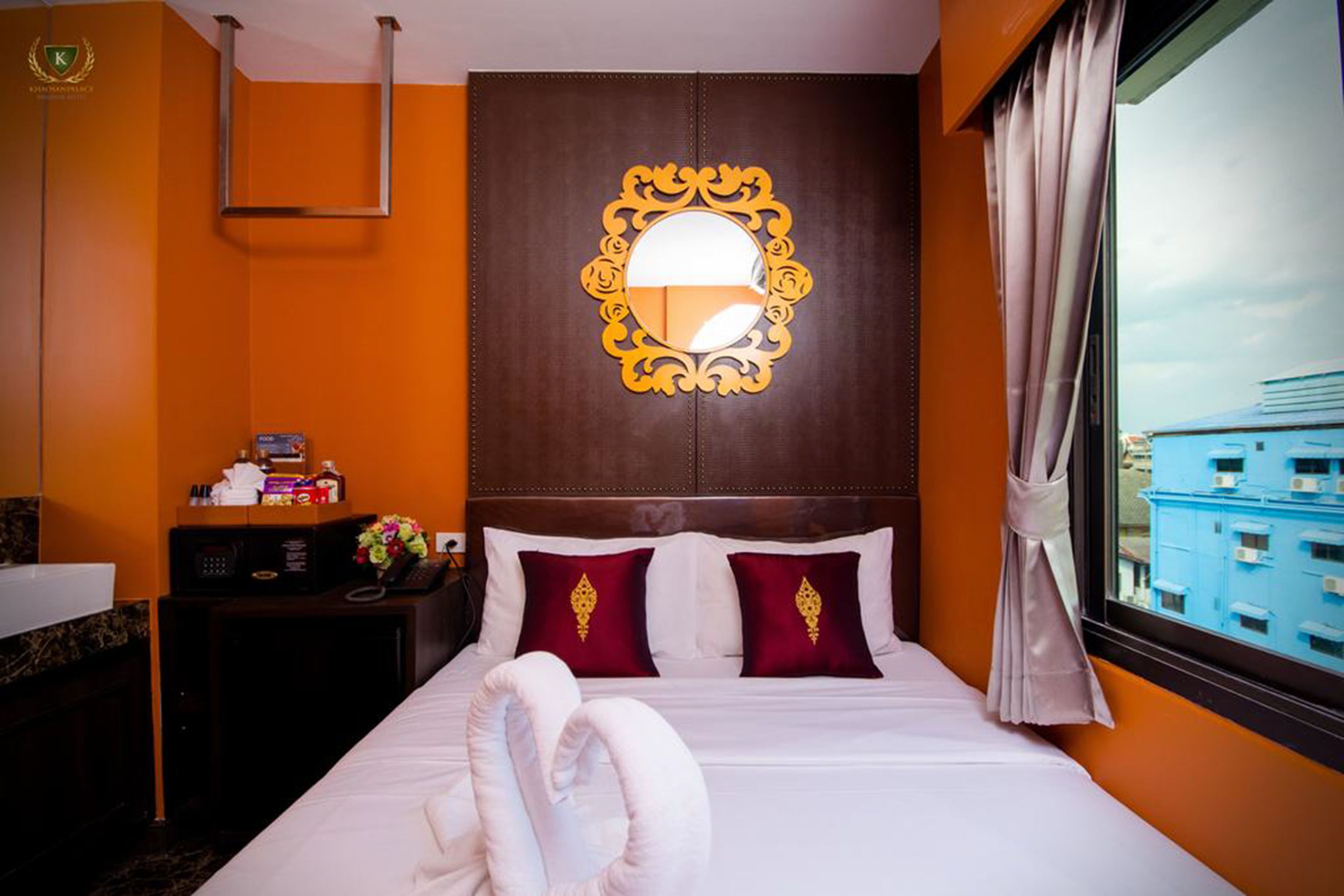 A hotel room with a bed, a headboard mirror, a bedside table with a phone and a box, and a window with curtains.