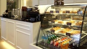 Um balcão de pequeno-almoço com uma vitrine de vidro que contém pastelaria e bebidas, painéis brancos nas paredes e uma máquina de café.