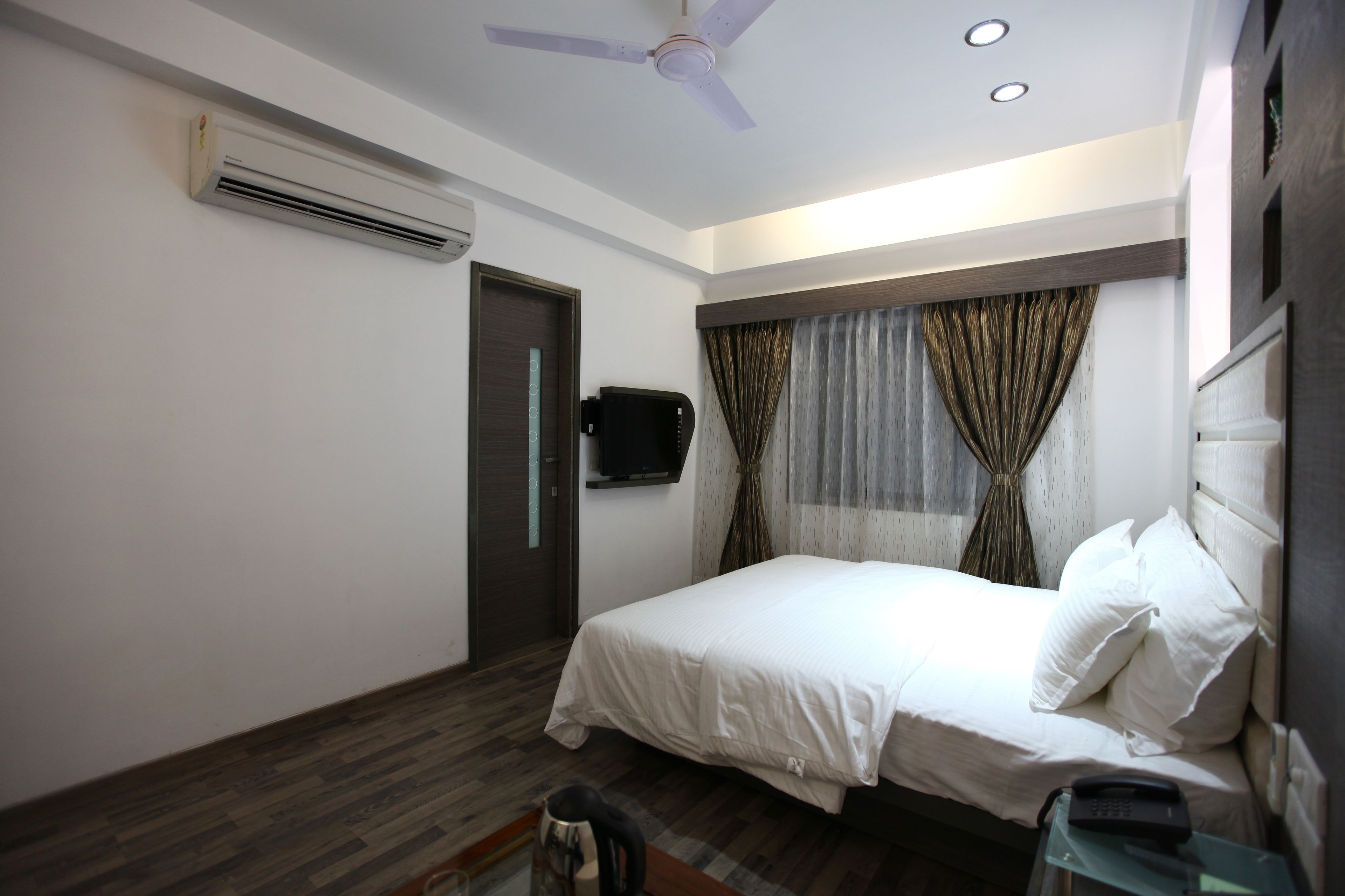 A bedroom with a bed, a ceiling fan, an air conditioning unit, a TV, and a bedside table.