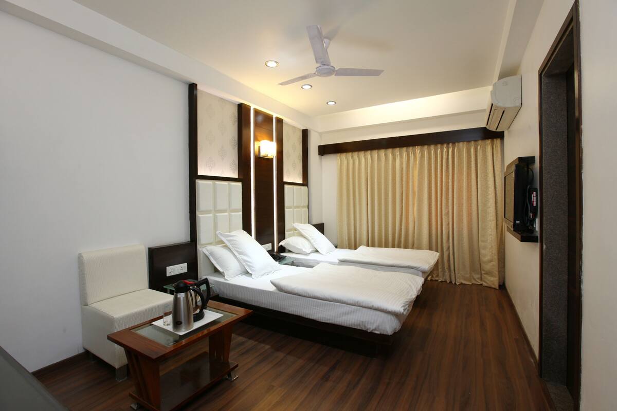 A hotel room with two single beds, a sofa, a small table with a kettle, a ceiling fan, and an air conditioning unit.