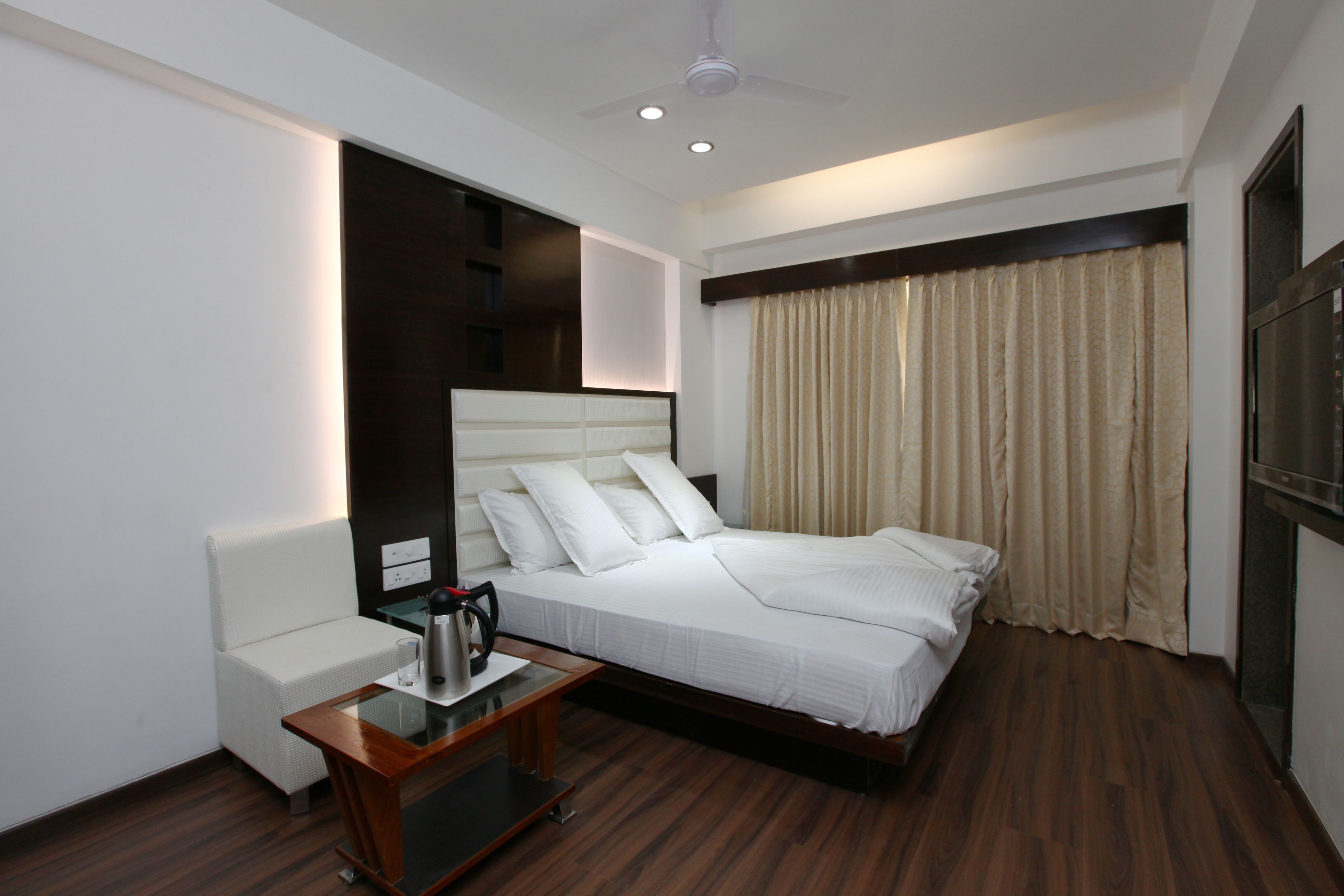 Una habitación de hotel moderna con una cama grande, una mesita de noche con hervidor, un sofá blanco y un ventilador de techo.