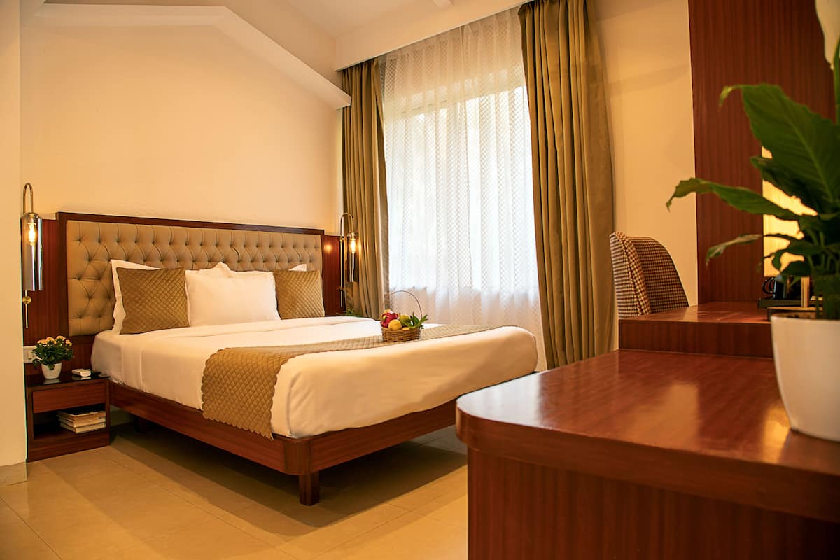 Een hotelkamer met een groot bed, een nachtkastje met een lamp, een klein tafeltje met een plant en een groot raam met gordijnen.